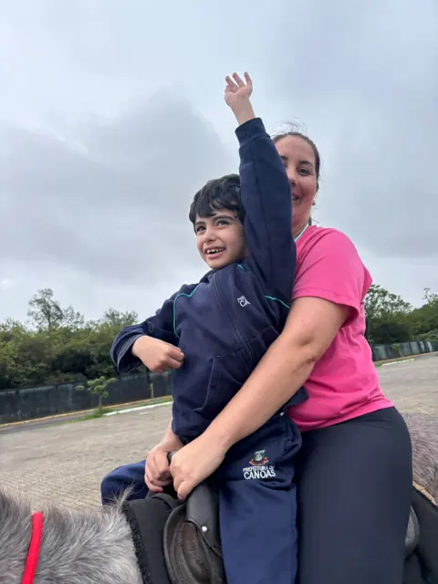 Criança com o braço erguido, sorrindo, montada com apoio de uma profissional