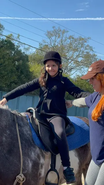 Menina de capacete montada, de braços abertos, com profissional ao lado
